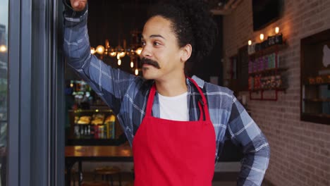 Mixed-race-male-barista-with-moustache-leaning-in-the-doorway-of-cafe-smiling