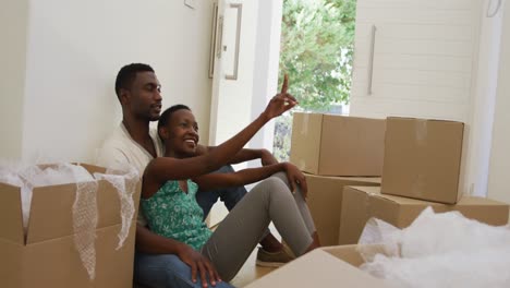 Happy-african-american-couple-embracing-and-talking-sitting-on-floor-with-packing-boxes