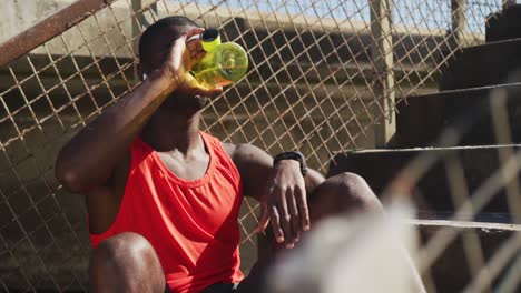Tired-african-american-man-sitting,-drinking-from-water-bottle,-taking-break-in-exercise-outdoors