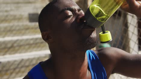 Tired-african-american-man-sitting-and-drinking-from-water-bottle,-taking-break-in-exercise-outdoors