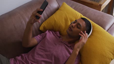 Mixed-race-man-wearing-headphones-lying-on-sofa-using-smartphone