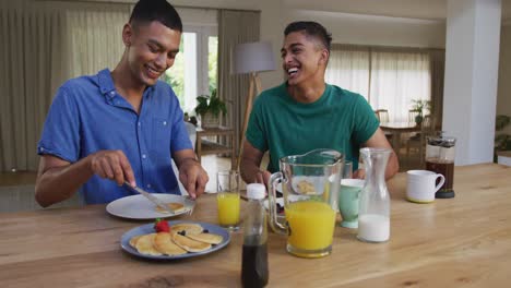 Happy-mixed-race-gay-male-couple-having-pancakes-for-breakfast-and-laughing-in-kitchen