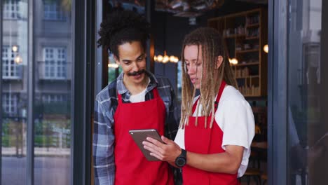 Two-diverse-male-baristas-wearing-aprons-standing-in-the-doorway-and-using-digital-tablet