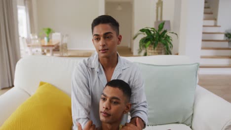 Smiling-mixed-race-gay-male-couple-sitting-in-living-room-holding-hands-embracing-and-talking