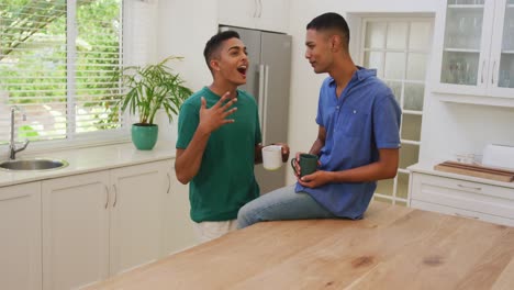 Happy-mixed-race-gay-male-couple-talking-and-drinking-coffee-in-kitchen