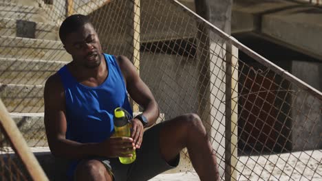 Tired-african-american-man-sitting,-holding-water-bottle,-taking-break-in-exercise-outdoors