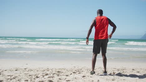 African-american-man-stretching-on-the-beach,-exercising-outdoors-by-the-sea