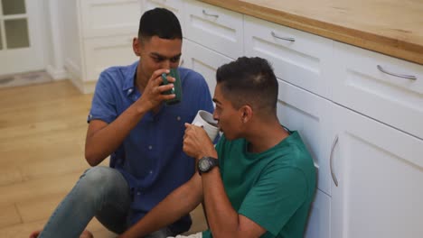 Happy-mixed-race-gay-male-couple-sitting-on-kitchen-floor-talking-and-drinking-coffee