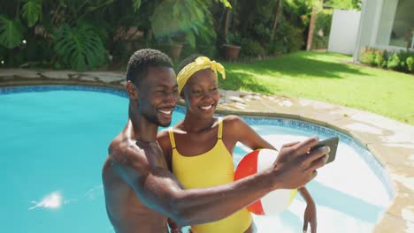 Happy-african-american-couple-standing-in-swimming-pool-taking-selfie-and-smiling-in-sunny-garden