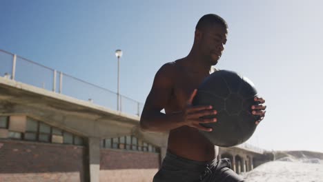 Focused-african-american-man-lifting-ball,-exercising-outdoors-on-beach