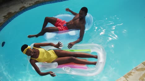 Glückliches-Afroamerikanisches-Paar,-Das-Auf-Schlauchbooten-Im-Schwimmbad-Liegt-Und-Sich-Gegenseitig-Bespritzt-Und-Lächelt