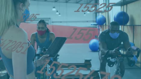 Animation-of-numbers-changing-over-female-instructor,-woman-and-man-in-face-masks-on-bikes-in-gym