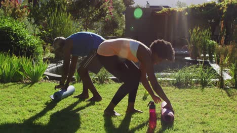 Afroamerikanisches-Seniorenpaar-Trainiert-Draußen-Und-Rollt-Yogamatten-Im-Sonnigen-Garten-Zusammen