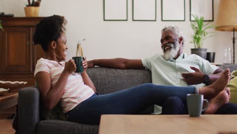 Glückliches-Afroamerikanisches-Paar-Sitzt-Auf-Der-Couch-Und-Redet,-Trinkt-Kaffee-Und-Lächelt