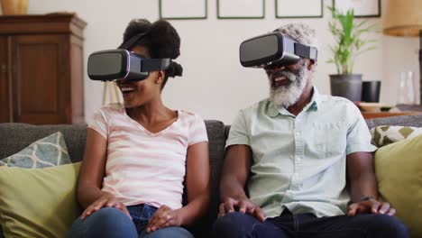 Happy-african-american-couple-sitting-on-couch-wearing-vr-headsets-and-smiling