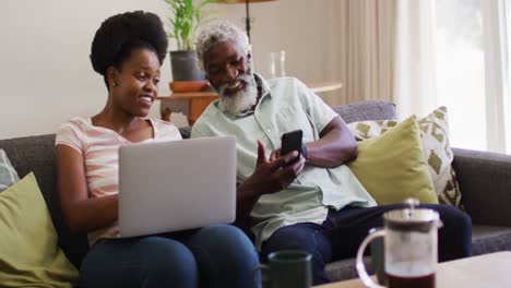 Happy-african-american-couple-sitting-on-sofa-talking-using-laptop-and-smartphone