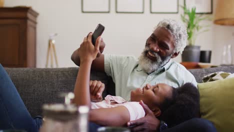 Happy-african-american-couple-relaxing-on-couch-looking-at-smartphone-and-smiling