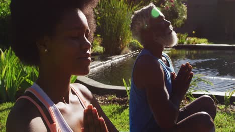 African-american-senior-couple-exercising-practicing-yoga-sitting-meditating-by-pond-in-sunny-garden