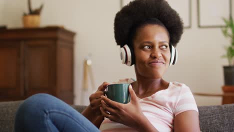 Feliz-Mujer-Afroamericana-Con-Auriculares-Relajándose-En-El-Sofá-Sosteniendo-Una-Taza-De-Café-Y-Sonriendo