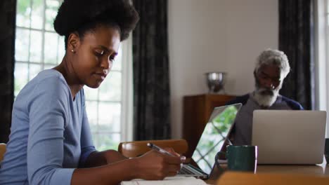 African-american-couple-sitting-at-dining-table-working-at-home-using-laptops