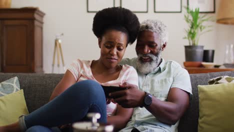 Happy-african-american-couple-sitting-on-couch-looking-at-smartphone-and-smiling