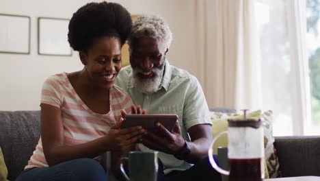 Happy-african-american-couple-sitting-on-couch-making-video-call-with-tablet-smiling-and-waving