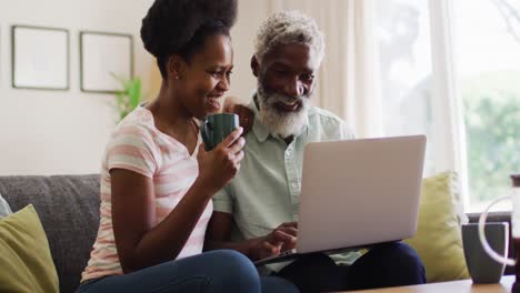 Happy-african-american-couple-sitting-on-sofa-smiling-and-using-laptop