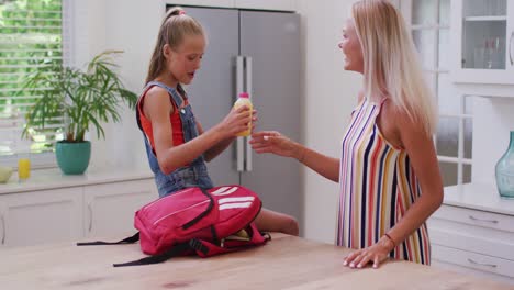 Smiling-caucasian-mother-in-kitchen-with-daughter-preparing-school-bag,-packing-juice-and-talking