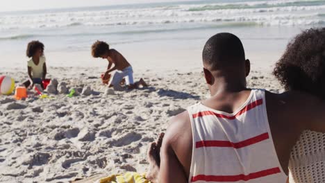 Afroamerikanische-Kinder-Spielen-Mit-Sand-Am-Strand,-Ihre-Eltern-Umarmen-Sich