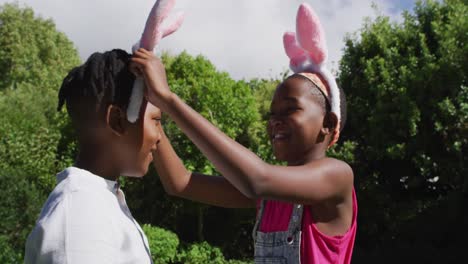 Lächelndes-Afroamerikanisches-Mädchen-Mit-Osterhasenohren,-Das-Ihrem-Bruder-Im-Garten-Ohren-Anlegt