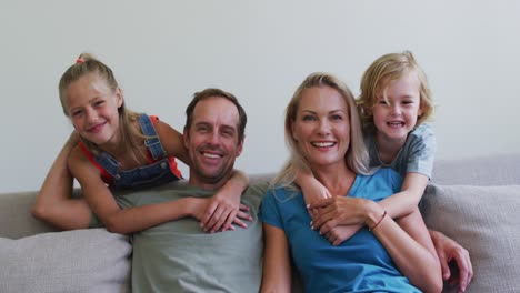 Portrait-of-smiling-caucasian-parents-on-sofa-with-son-and-daughter-embracing-them