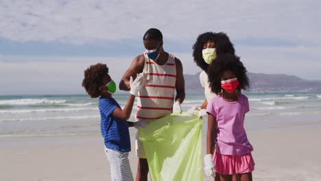 Afroamerikanische-Eltern-Mit-Zwei-Kindern,-Die-Gesichtsmasken-Tragen,-Sammeln-Müll-Vom-Strand