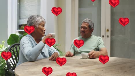 Animation-of-red-hearts-over-senior-couple-drinking-coffee-at-home