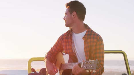 Happy-caucasian-man-sitting-in-beach-buggy-by-the-sea-playing-guitar