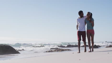 African-american-couple-smiling,-embracing-and-walking-on-the-beach