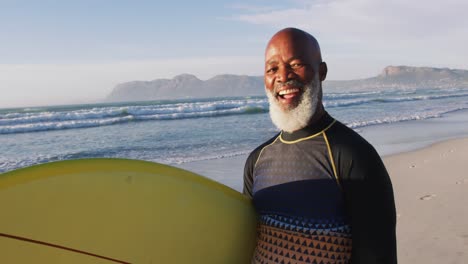 Lächelnder-älterer-Afroamerikanischer-Mann,-Der-Mit-Einem-Surfbrett-Am-Strand-Spaziert