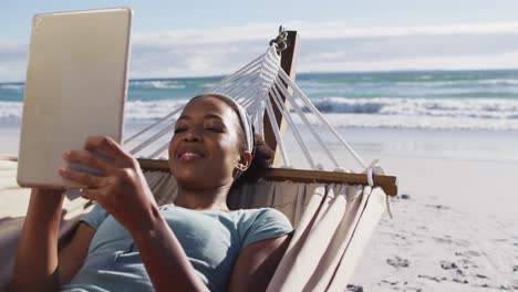 Afroamerikanische-Frau-Benutzt-Digitales-Tablet,-Während-Sie-Auf-Einer-Hängematte-Am-Strand-Liegt