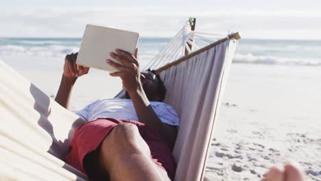 African-american-man-using-digital-tablet-while-lying-on-a-hammock-at-the-beach