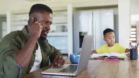 Afroamerikanischer-Vater-Und-Sohn-Machen-Hausaufgaben,-Reden-Auf-Dem-Smartphone-Und-Benutzen-Gemeinsam-Einen-Laptop