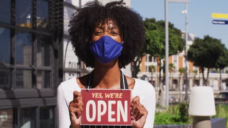 African-american-female-cafe-worker-wearing-face-mask-showing-were-open-signage