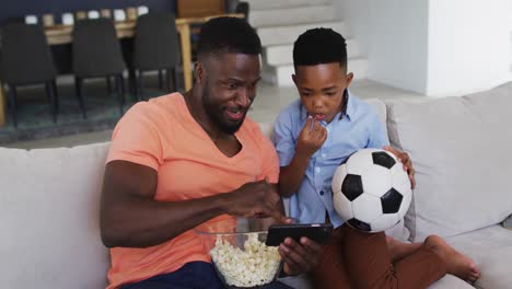 Afroamerikanischer-Vater-Und-Sohn-Essen-Popcorn-Und-Schauen-Sich-Ein-Fußballspiel-Auf-Dem-Smartphone-An