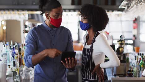 Diverse-male-and-female-cafe-workers-wearing-face-masks-in-cafe-using-digital-tablet-talking
