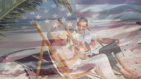 Amerikanische-Flagge-Winkt-Gegen-Ein-älteres-Paar,-Das-Getränke-Genießt,-Die-Auf-Einem-Liegestuhl-Am-Strand-Sitzen