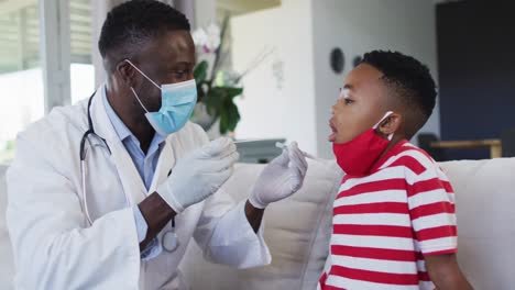Afroamerikanischer-Arzt-Mit-Gesichtsmaske,-Der-Zu-Hause-Eine-Rachenabstrichprobe-Eines-Jungen-Entnimmt