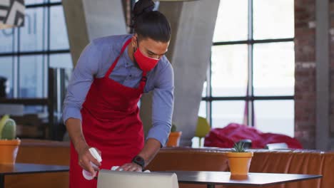 Mixed-race-male-cafe-worker-wearing-face-mask-disinfecting-table