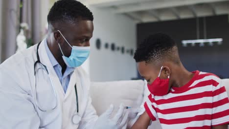 Médico-Afroamericano-Con-Mascarilla-Inyectando-La-Vacuna-Covid-19-A-Un-Niño-En-Casa