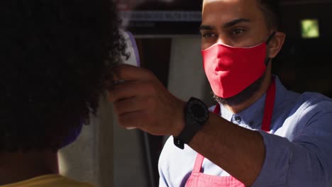 Mixed-race-male-cafe-worker-wearing-face-mask-taking-temperature-of-female-coworker