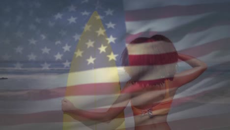 Amerikanische-Flagge-Weht-Vor-Der-Rückansicht-Einer-Frau-Mit-Surfbrett,-Die-Am-Strand-Steht