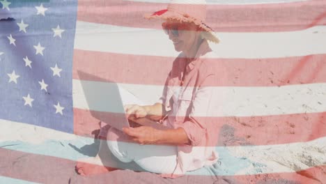 Amerikanische-Flagge-Winkt-Gegen-Eine-ältere-Kaukasische-Frau,-Die-Einen-Laptop-Benutzt,-Während-Sie-Am-Strand-Sitzt
