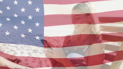 Animation-of-american-flag-waving-over-smiling-woman-in-deckchair-using-smartphone-on-beach